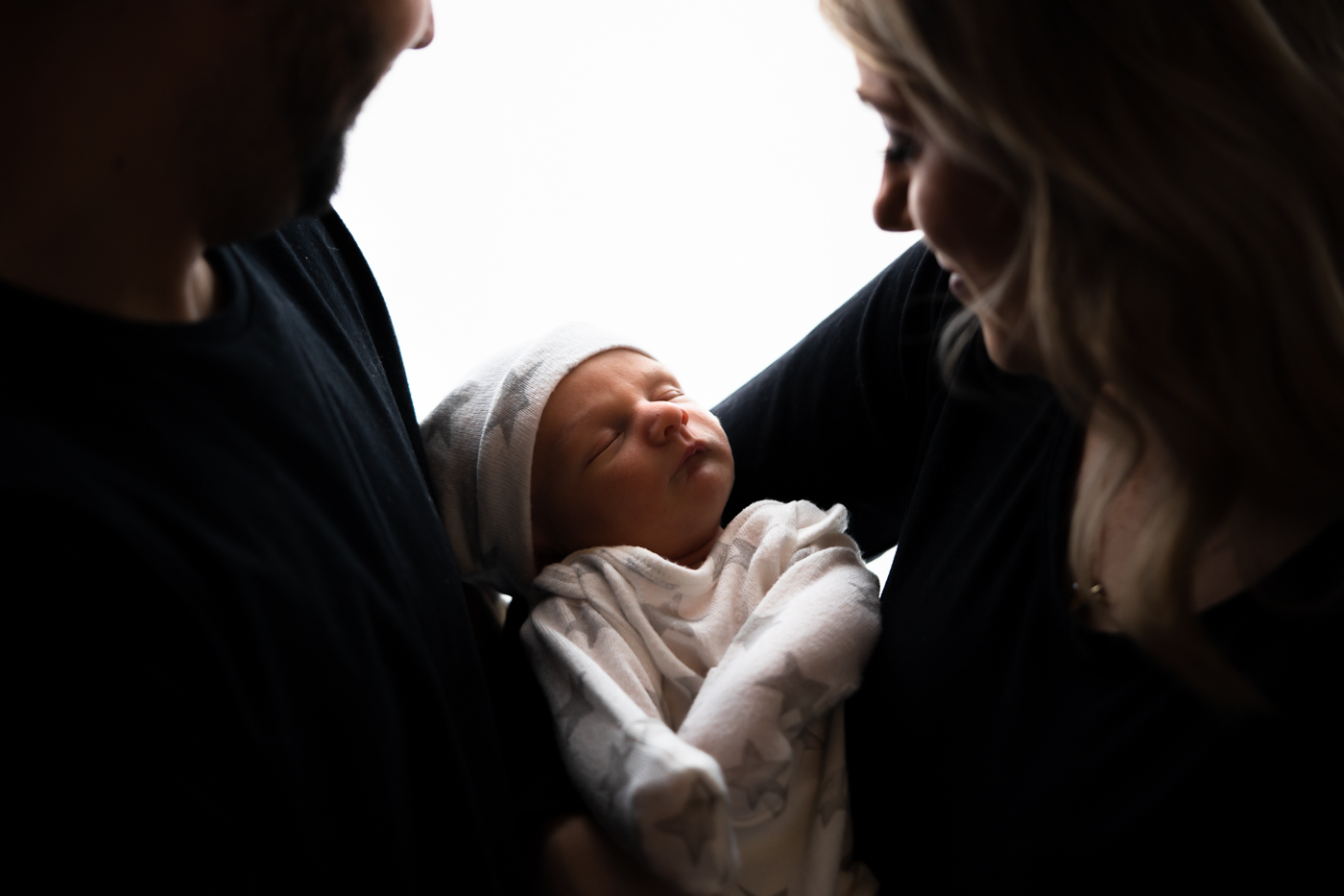 A close-up photograph of a mother and father holding their newborn baby boy in front of a white window. The parents are looking down at the baby with love and tenderness. The baby is wrapped in a blanket and is sleeping peacefully. The window is letting in natural light, which illuminates the scene.