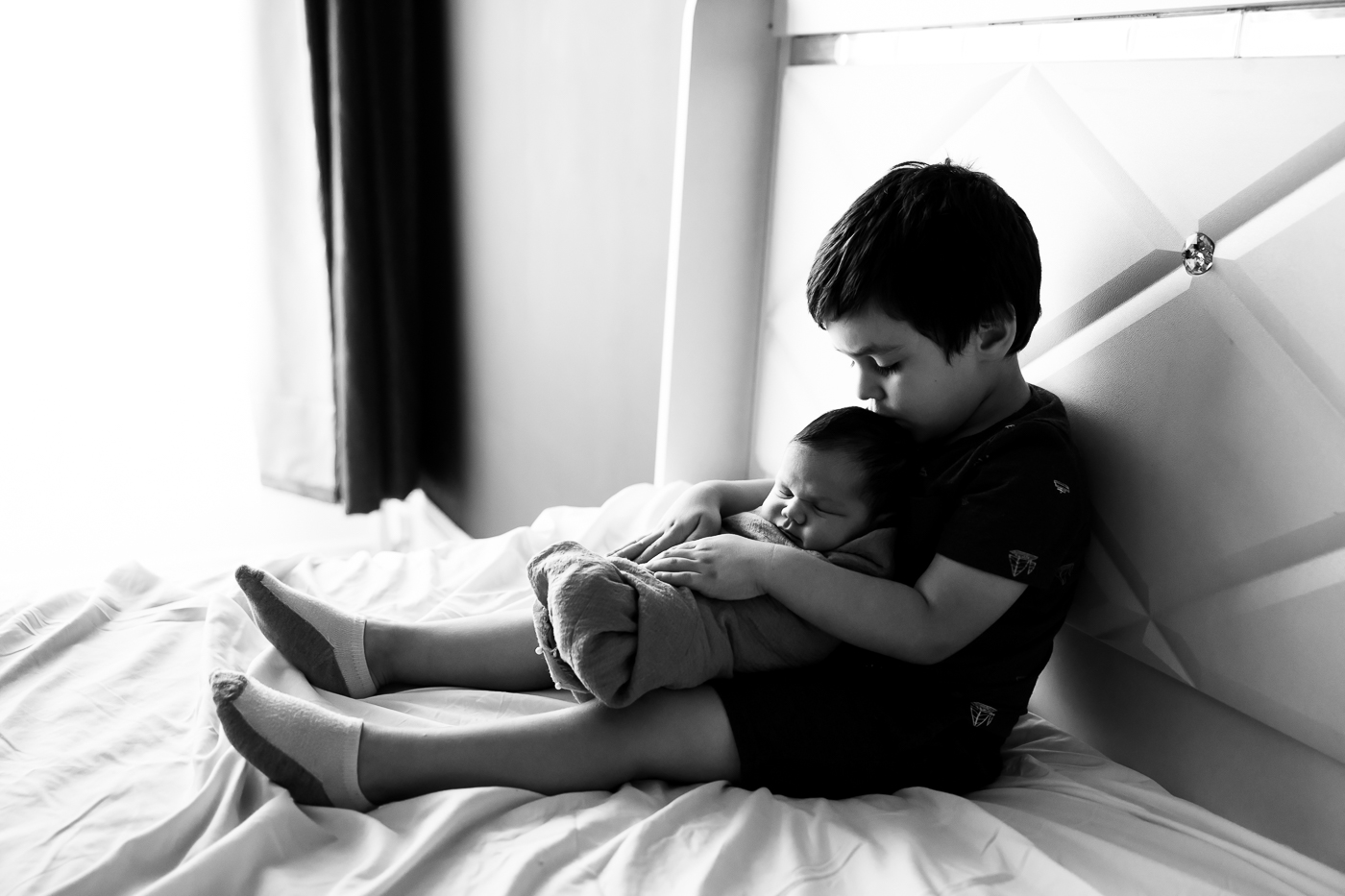 Black and white image of a toddler holding his baby sister in his arms and kissing the top of her head. The baby is asleep. The image conveys a sense of love, affection, and protection.