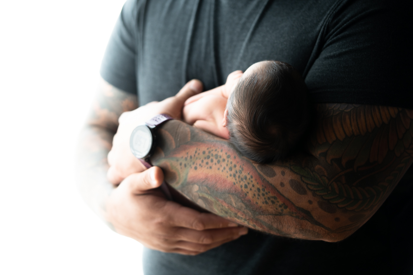 Close-up of a newborn baby girl's head resting on her father's colorfully tattooed arms.