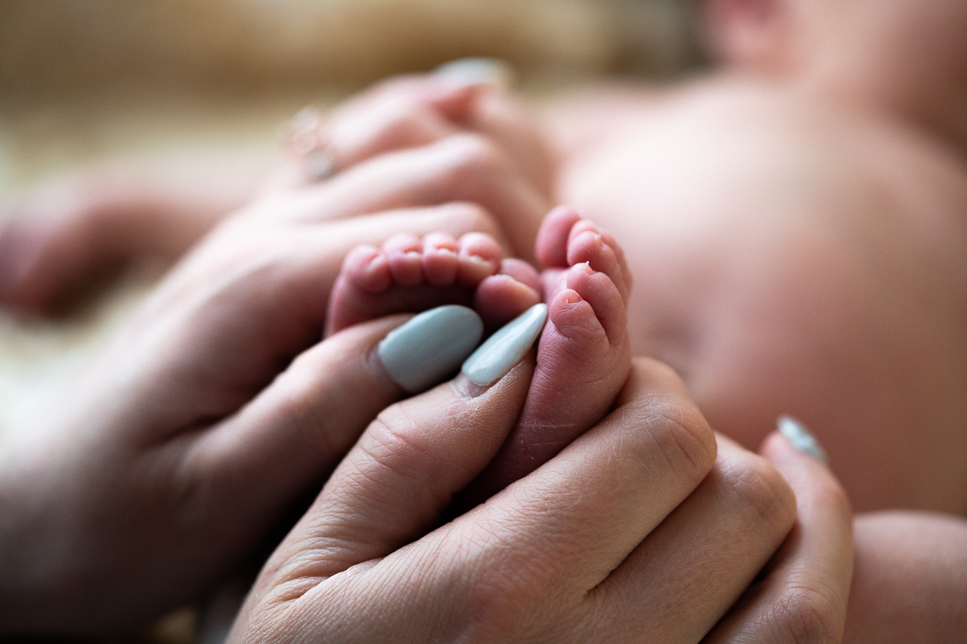 Close-up photograph of a mother's hands holding her baby's feet. The mother's hands are gentle and loving, and the baby's feet are tiny and delicate. The image conveys a sense of warmth, love, and protection.